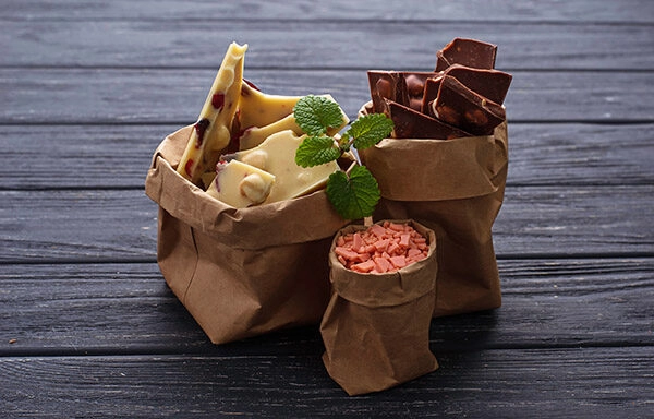 Bolsa de papel con diferentes tipos de chocolate, incluyendo chocolate blanco, chocolate con frutas y chocolate oscuro. Complemento de chocolate en trozos y decorado con una ramita de menta fresca. Fondo de madera oscura que resalta los colores de los chocolates. Ideal para amantes del dulce y la repostería.