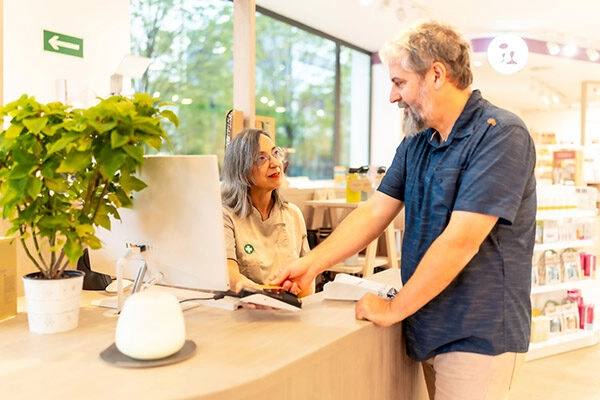Cliente interactuando con una empleada en un mostrador de atención al cliente en una tienda. La mujer sonriente ofrece un servicio amable mientras el hombre está en proceso de realizar una compra. El ambiente es moderno y luminoso, con estanterías de productos visibles al fondo. Ideal para promocionar atención al cliente y experiencias de compra positivas.