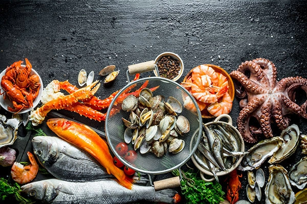 Variedad de mariscos frescos dispuestos sobre una superficie oscura, incluyendo camarones, pulpo, cangrejo, almejas y diferentes tipos de pescado. La imagen muestra un colador con mariscos, resaltando la frescura y la diversidad de productos del mar, ideal para recetas de cocina.